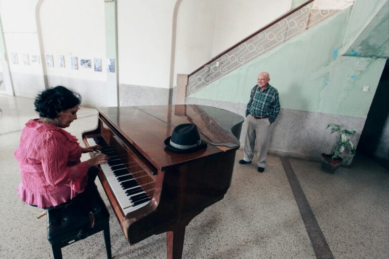A woman plays a grand piano inside a rustic room with an observer nearby, creating an intimate musical setting.